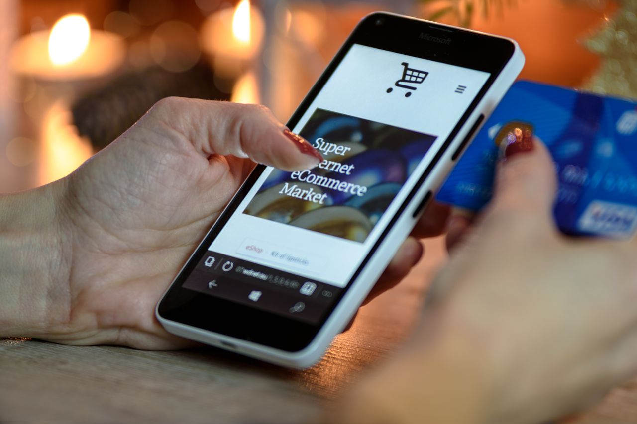 home-hero-img Woman using a smartphone for online shopping, holding a credit card in hand, with festive lighting in the background.